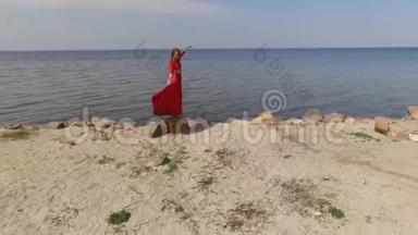 穿着红色长裙，手舞足蹈的娇艳女子举起手在蓝色海水的壮观景色附近的大石头上