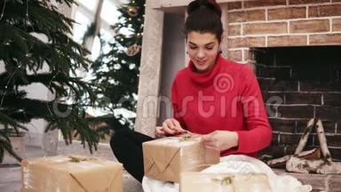 美丽的微笑女人把圣诞礼物包在家中的圣诞树和壁炉旁，然后把礼物放在