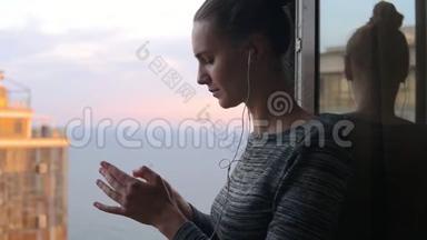 在阳光明媚的一天里，微笑的女孩站在窗边，用手机耳机听音乐