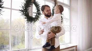 年轻的父亲抱着他的小男孩，站在窗边看着外面。 窗户上装饰着圣诞节