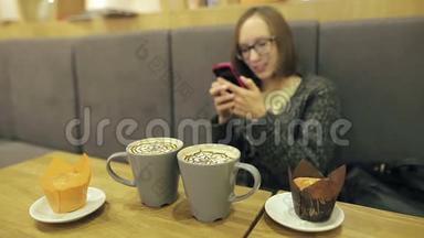 戴眼镜的女人在咖啡馆里使用智能手机上的应用程序，喝椰子咖啡，微笑着，在手机上发短信。 美丽美丽