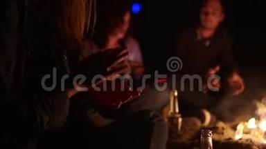 两个男孩和两个女孩晚上坐在炉火旁唱歌。 快乐的朋友唱歌弹吉他