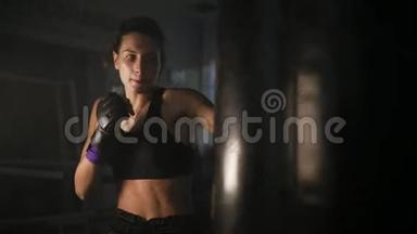 漂亮的女踢脚手在黑暗的健身工作室训练时，用她的拳头和肘部<strong>打拳击</strong>袋。 凶猛
