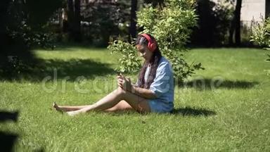 小女孩听音乐，戴着耳机坐在公园的草地上。 4K