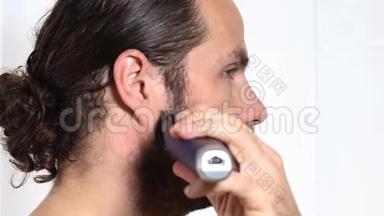 英俊的年轻人留着长发，在浴室里用修剪器刮胡子。 特写镜头。