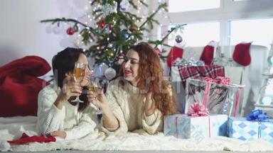 年轻漂亮的黑发女人和红发女人躺在靠近圣诞杉树的地毯上，戴着叮当作响的眼镜，喝着香槟