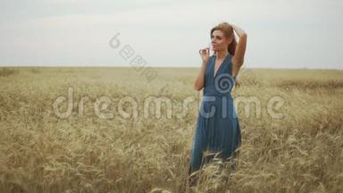美丽的年轻女子穿着一件蓝色长裙，站在金色的麦田里，试着`麦茎，抚摸着她的头发，抚摸着她的头发