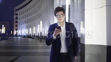 年轻的商人带着智能手机和耳机走在城市的一条夜晚街道上