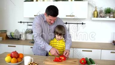 父亲和儿子在家厨房里切蔬菜。 父亲协助非常可爱的儿子在厨房切蔬菜。 切割