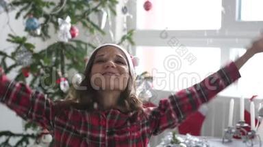 戴着圣诞帽的快乐年轻漂亮的女人把纸屑扔在窗边的圣诞树旁，并展示了它的形状。