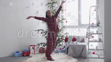 穿着睡衣的年轻黑发女人站在圣诞树附近，把纸屑扔到窗边。 慢动作