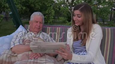 一个年轻的女孩帮助一个坐在秋千上的生病的老人读报纸。