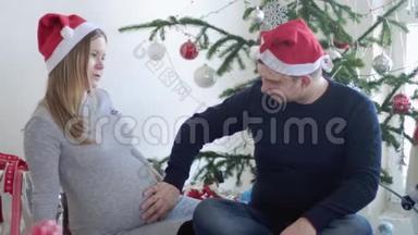 快乐的年轻家庭。 丈夫拥抱他怀孕的妻子，抚摸她的腹部附近美丽的<strong>装饰</strong>圣诞树。 慢动作