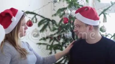 戴着圣诞帽的幸福年轻夫妇正在美丽的圣诞树旁接吻。 慢动作。 关门