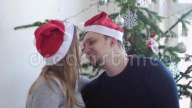 圣诞老人帽里快乐的年轻家庭。 丈夫亲吻他怀孕的妻子，靠近美丽的<strong>装饰</strong>圣诞树。 slwo运动