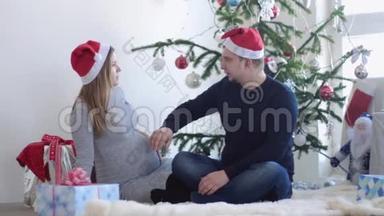 挺年轻的。 丈夫抚摸他怀孕的妻子，靠近美丽的装饰圣诞树。 慢动作