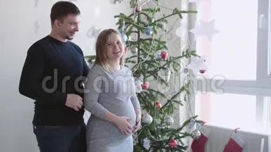 年轻快乐的家庭。 丈夫在画室窗户旁美丽的圣诞树旁拥抱他怀孕的妻子