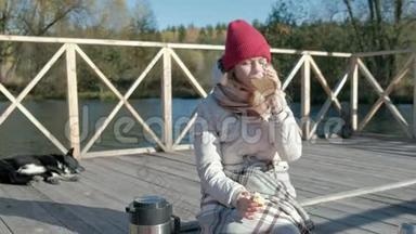 一位穿着<strong>保暖</strong>衣服的女游客在河岸附近的一座桥上背着背包，喝着热茶，野餐，一只狗躺着