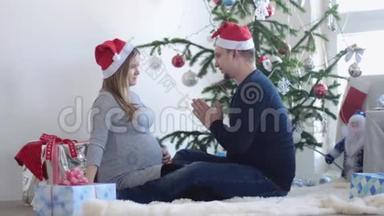 戴着圣诞帽的幸福年轻夫妇坐在靠窗的漂亮圣诞树旁的地毯上。 慢动作