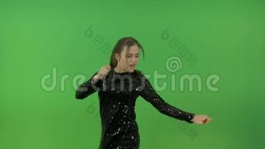 一个穿着黑色闪亮连衣裙，跳舞唱歌的年轻女孩。 欢快的跳跃和欢乐。 在绿色的背景上。