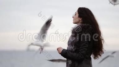一个年轻漂亮的黑发女人的肖像，穿着毛皮大衣，冬天在海边喂海鸥。慢镜头