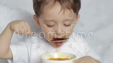 这个男孩高兴地用勺子从盘子里吃汤，<strong>特写</strong>。 运动相机滑块。 一个男孩坐在那儿<strong>吃饭</strong>