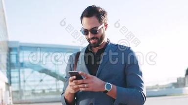 一位留着大胡子的时髦男士站在机场候机楼旁，用手机旋转着，迷人地微笑着。