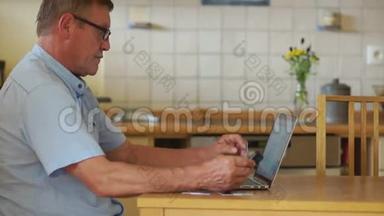 一位老人拿着电脑和电话工作，坐在厨房的桌子旁。 视力和<strong>防</strong>护<strong>眼镜</strong>