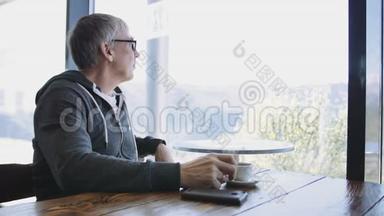 白种人高级运动风格的男人看着窗外的咖啡馆。 一个年老的灰色男人在桌旁喝咖啡