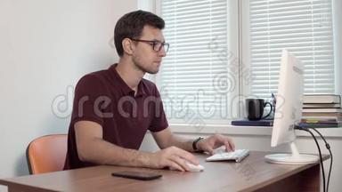 年轻的办公室工作人员戴着眼镜坐在窗边的办公室里，用电脑打字报告。 智能手机