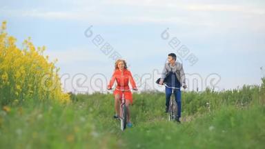 正面看着美丽欢快的情侣们积极骑着自行车沿着绿色盛开的田野。