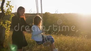 快乐的年轻妈妈和一个小女孩在日落时一起在公园里摇摆。 妈妈和女儿骑着秋千，家人