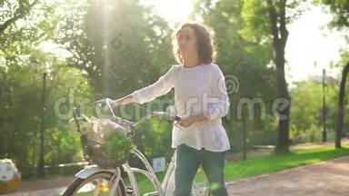 穿着白色衬衫和蓝色牛仔裤的漂亮女人拿着她的城市自行车走在城市公园的小巷`