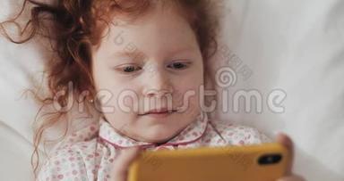 有趣的红发小女孩躺在床上，饶有兴致地在现代黄色智能手机上观看视频。