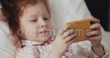 有趣的红发小女孩躺在床上，饶有兴致地在现代黄色智能手机上<strong>观看视频</strong>。