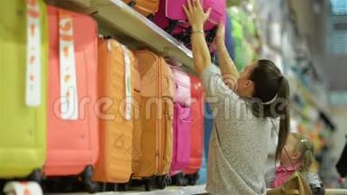 布鲁内特母亲和她的小女儿选择旅行袋。 可爱的女孩坐在超市的购物车里，年轻的女人