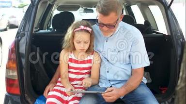 孙女和祖父坐在汽<strong>车后备箱</strong>里，翻阅着平板电脑里的照片。家庭休闲