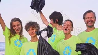 5名年轻志愿者身穿绿色t恤衫，将图像回收，在海洋海滩上收集垃圾，然后在相机上摆姿势