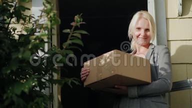 一个拿着包裹的女人站在她家的门槛上。 看着相机，微笑着。 将包裹送达