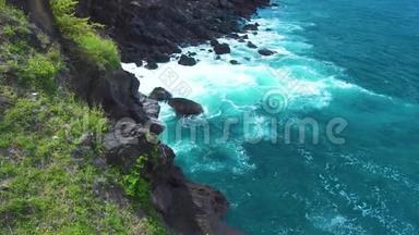 在绿色植物的背景下，海浪冲破岩石悬崖。 汹涌的海浪向岩石岛溅起泡沫