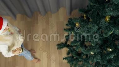 慢动作俯视镜头.. 小女孩把金球挂在圣诞树上。 穿红色圣诞帽的女孩装饰圣诞树