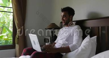 西班牙裔男子使用笔记本电脑坐在床上，在卧室里上网<strong>聊天</strong>，在热带景色中寻找<strong>窗口</strong>