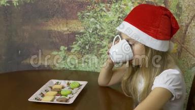 戴着红帽子的白种人年轻女孩坐在家庭厨房的桌子上。 女孩喝水或茶从白色杯子。 关门