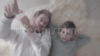有趣的有胡子的爸爸和他的小儿子躺在白色的毛皮地毯上，有有趣的谈话。 父亲-子女
