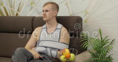 一个痛苦的男人，<strong>胳膊</strong>断了，戴着手臂支撑，坐在沙发上看电视