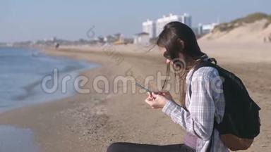 旅行者女孩带着背包坐在沙滩上，在手机上输入一条信息。