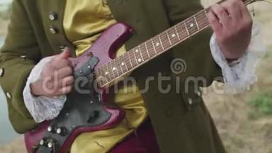 为一个穿着洛可可式老式服装在枯草上演奏的紫色电吉他的男人靠近