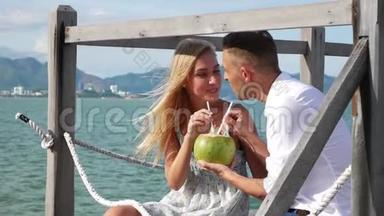 年轻的幸福夫妇坐在木墩上，笑着喝着蓝色海洋背景上的椰子。 浪漫浪漫
