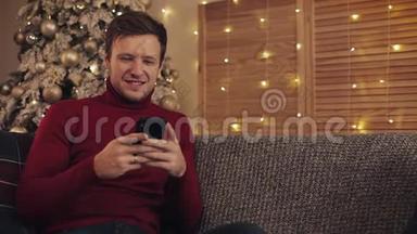 迷人的男人使用智能手机坐在沙发上，发信息，微笑在装饰好的公寓附近。 圣诞树