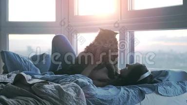 戴着耳机的年轻美女在床上放松，她可爱的缅因州浣熊猫在窗边听音乐